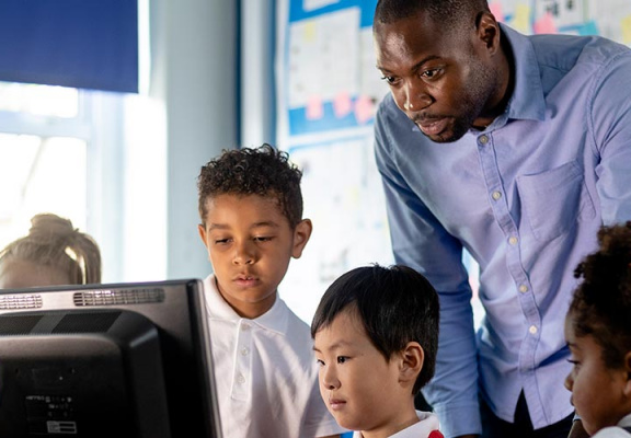 A low-angle view of a teacher and his students learning in an ICT classroom suite. The little boy in red is sat at the desk showing the group his work on the computer.